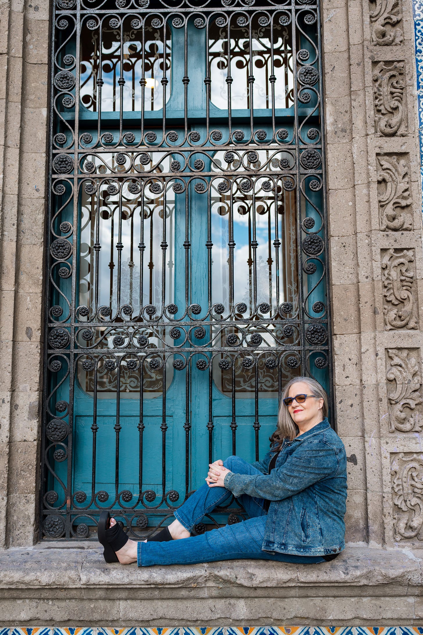 Me sitting cross-legged in front of a tall, ornate turquoise door framed by carved stone and wrought iron in Mexico City. I'm wearing sunglasses, a denim jacket, and jeans, looking relaxed and reflective.