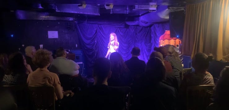 small woman with ginger hair and white tshirt stands on stage with a mic. Audience sit facing her