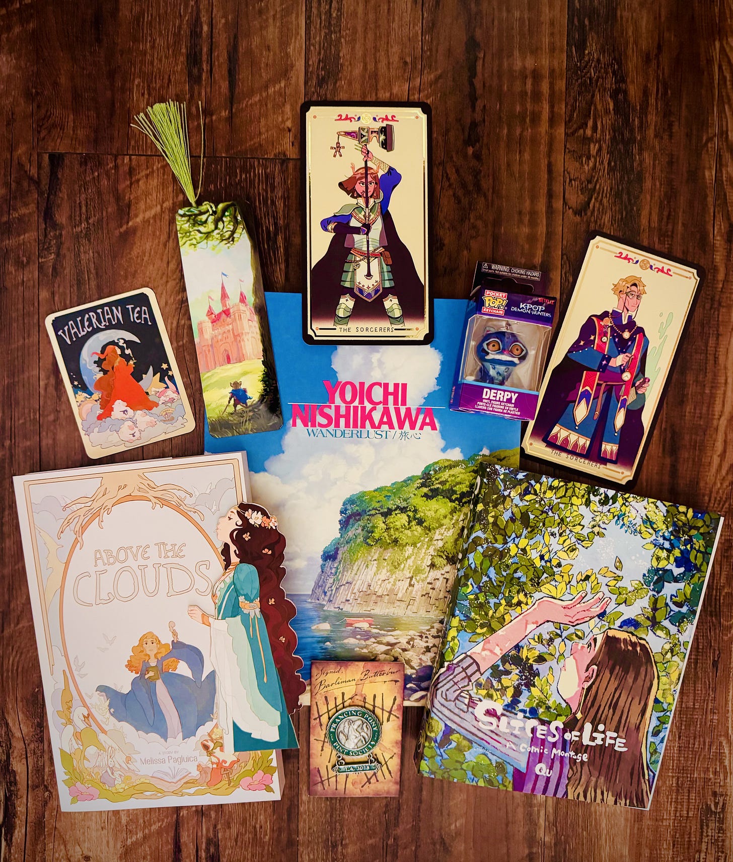 Photos of book covers of Wanderlust by Yoichi Nishkawa, Slices of Life by Qu, and Above the Clouds by Melissa Pagluica. The photo also contains fantasy and brightly colored bookmarks, art cards, and stickers as well as a figurine of Derpy, the blue tiger from Kpop Demon Hunters.
