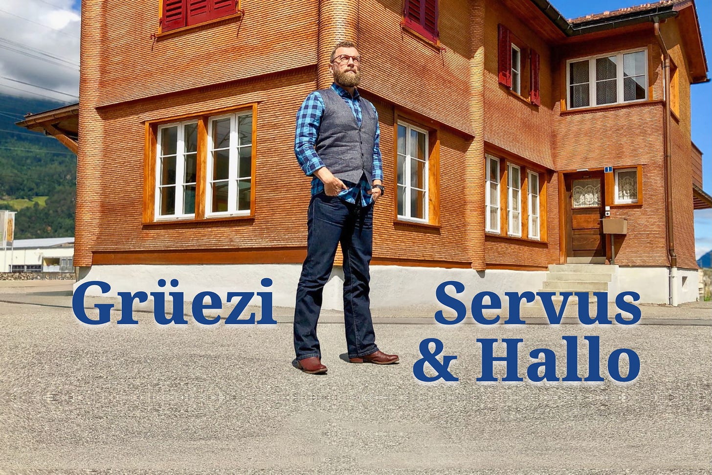 Jürg Vollmer steht vor dem alten Stationsgebäude des Bahnhofs Zizers. Er trägt Jeans, kariertes Hemd und Weste. Das Gebäude hinter ihm ist mit feinen, horizontalen Holzschindeln verkleidet, hat rote Fensterläden und Fenster mit weissen Rahmen. Der Himmel ist klar blau, und die Szene wirkt sonnig und ruhig. Unten am Bild steht die Begrüssung: Grüezi, Servus und Hallo.