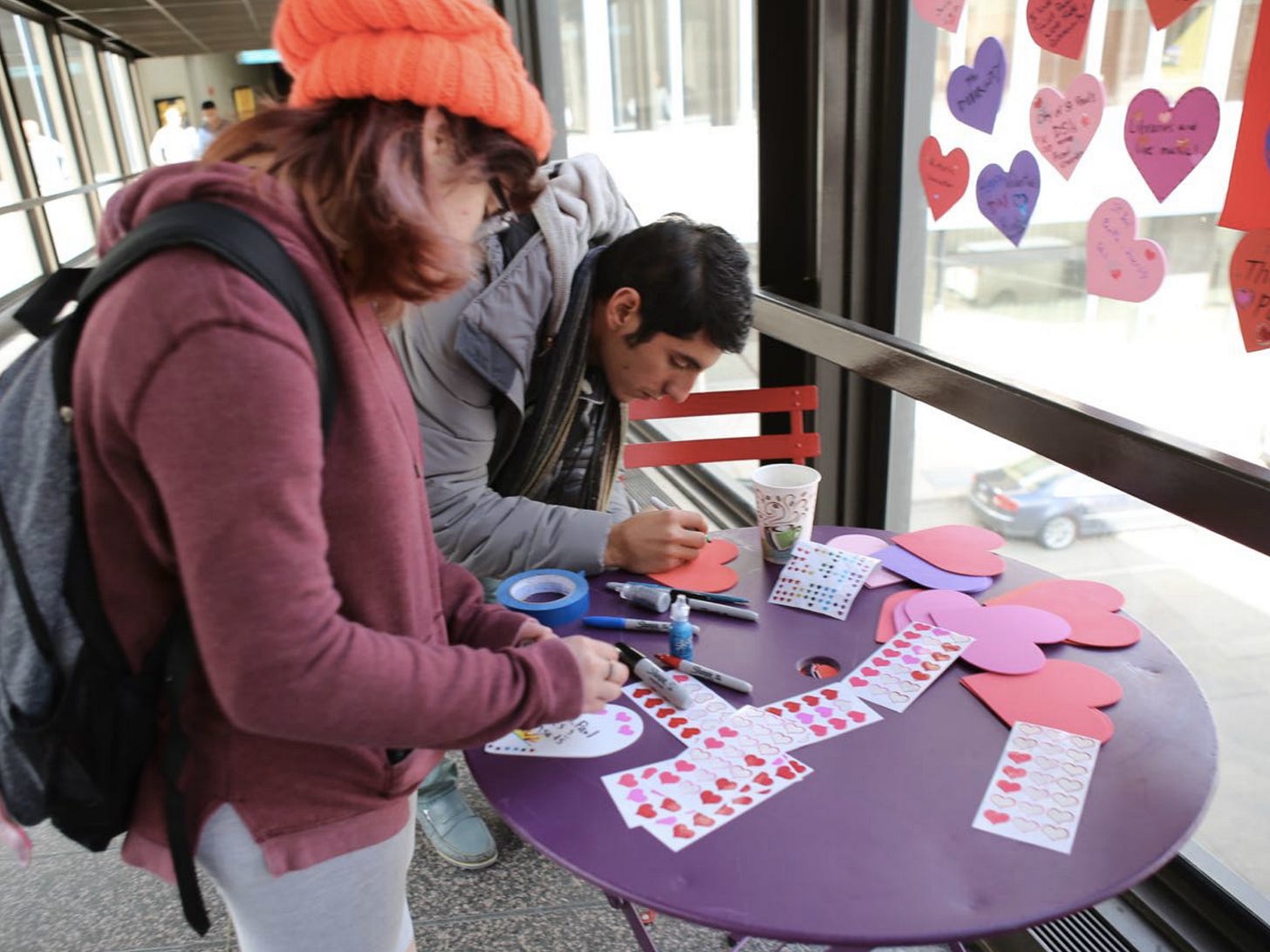 Two young people create Valentine's Day cards at a table in a skyway. On the window next to them are many more heart-shaped cards that other people have made.