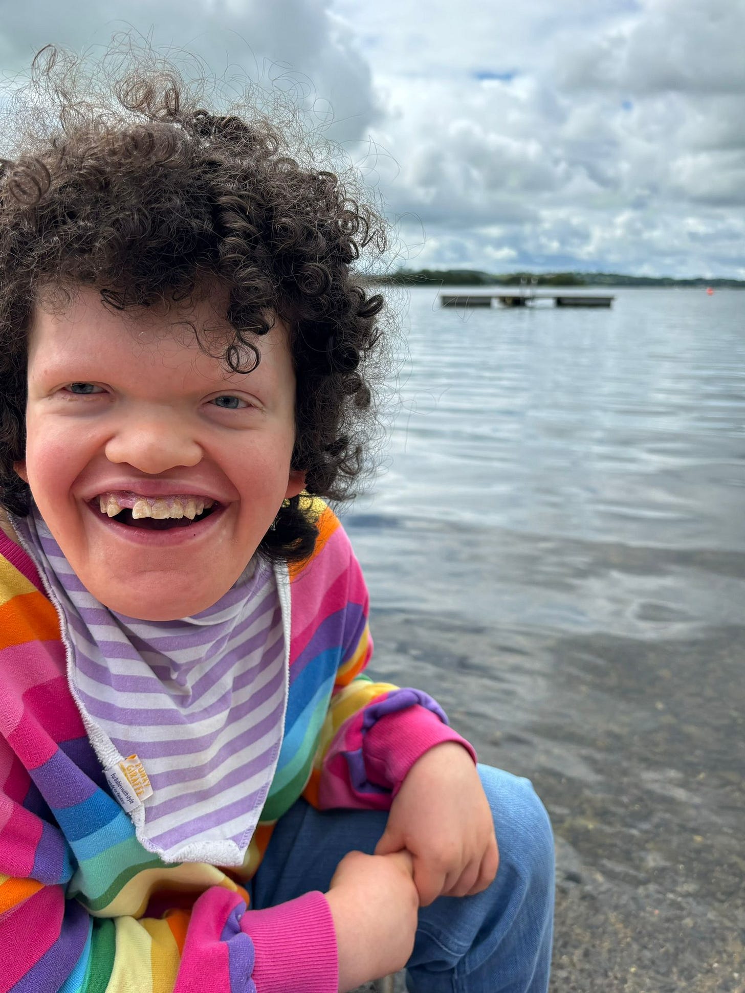 Smiling girl with blue eyes and dark curly hair wearing a rainbow bright sweatshirt on a grey day with a cloudy sky and lake in the background