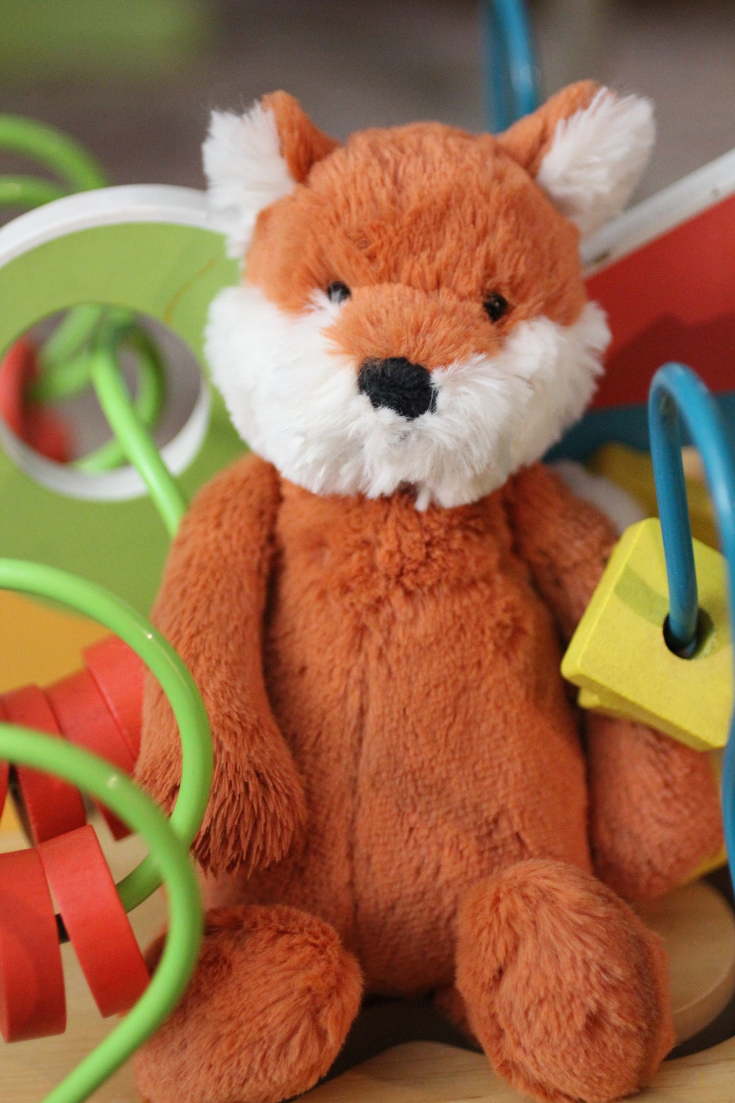 Plush toy fox with orange fur and white face sitting among colorful children’s toys and activity beads.
