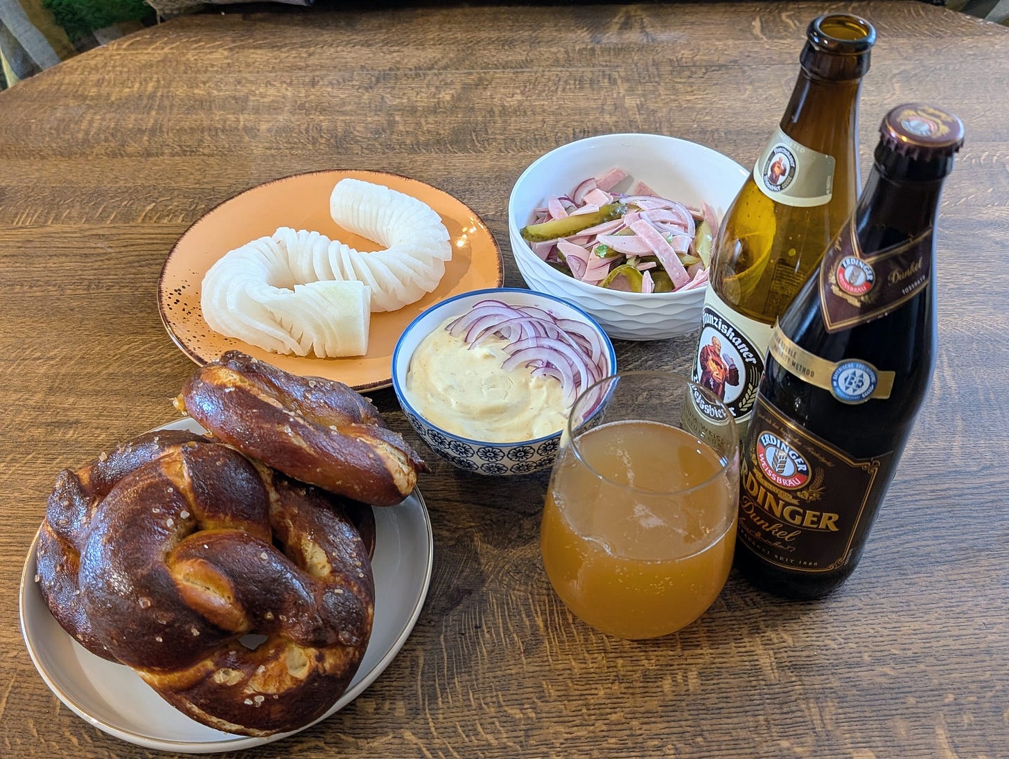 A wooden table set with a Bavarian-style spread: two large glossy pretzels on a plate, a bowl of orange‑tinted Obatzda topped with thin slices of red onion, a plate of neatly spiralled salted white radish, and a bowl of Wurstsalat made from sliced sausage and pickles. Beside the food are two German beer bottles—Franziskaner Weissbier and Erdinger Dunkel—and a glass filled with pale amber beer.