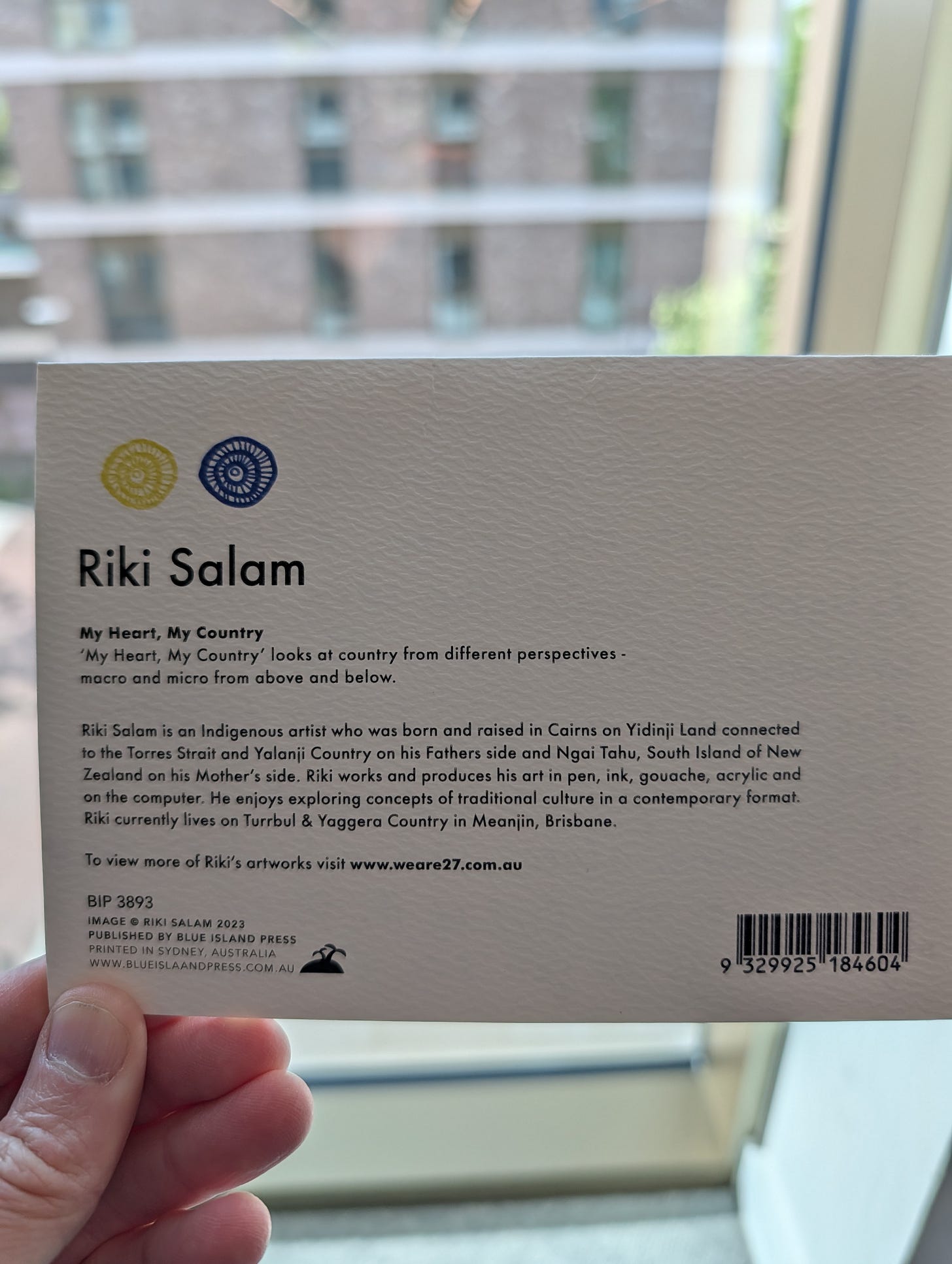 Back of a white card showing gold and navy circular logos and text about Riki Salam, an Indigenous artist from Brisbane. The card reads "My Heart, My Country" and includes artist bio, publisher details (Blue Island Press, Sydney, 2023), and barcode.