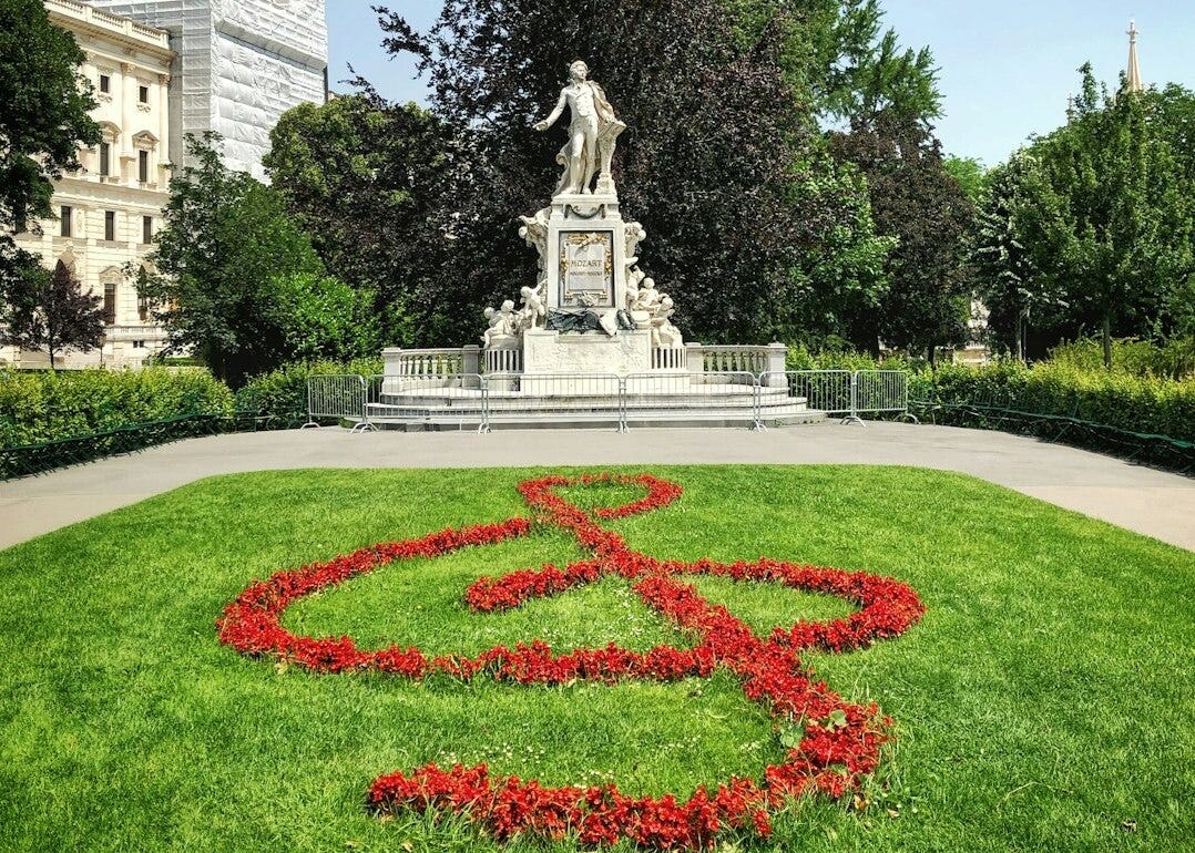 Estatua de Mozart em um jardim, que na frente tem um gramado verde e uma clave de sol vermelha, formada aparentemente por flores do jardim.