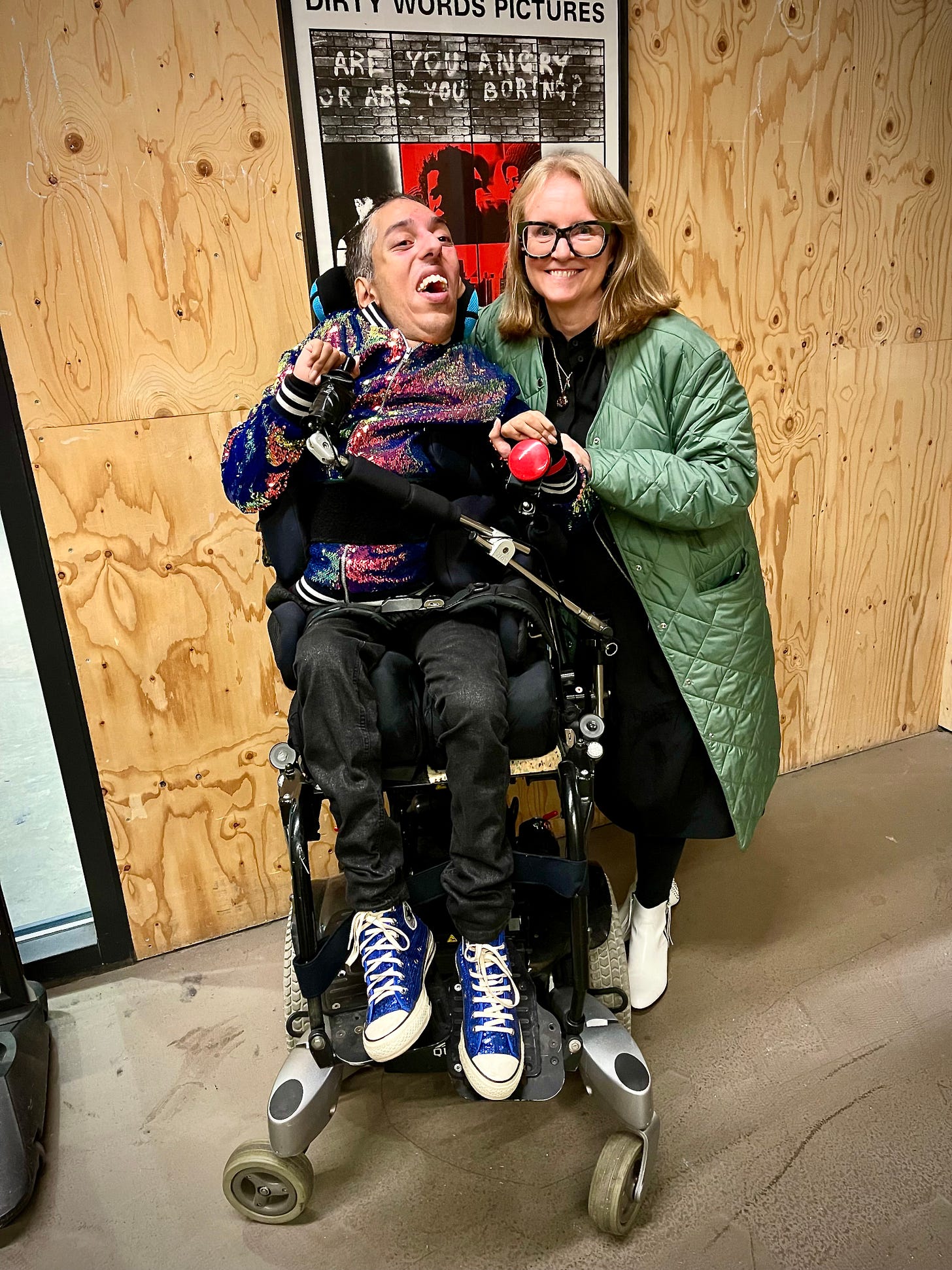 Image Description 1: In front of a film poster on a wooden wall at Central Saint Martins, Sulaiman (a wholeheartedly Disabled AF and British-Pakistani man with black buzzcut hair and features “white hair of wisdom”), sits in his power wheelchair next his friend Deborah (a badass white woman with shoulder-length blonde hair and large black framed glasses. The top of the film poster can be seen just over their heads and is of white spray painted all caps words on an image of a stone wall saying, “Are you angry or are you boring?” Sulaiman is wearing his joyous sequinned casual jacket, his black skinny jeans with silver metallic thread throughout, and his blue sequinned converse shoes. Deborah is wearing a knee-length pale green down coat, a long black dress, black tights, and white boots. Both, Sulaiman and Deborah, are smiling and joyful. END. 
