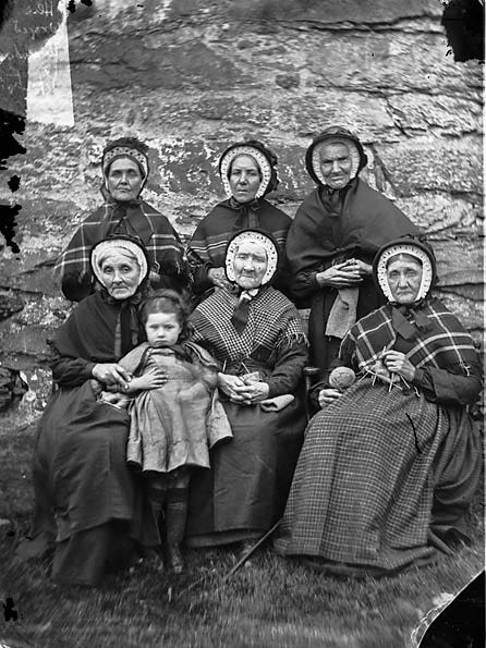 Black and white photograph of six elderly women and a young child posed against a stone wall, one woman knitting.
