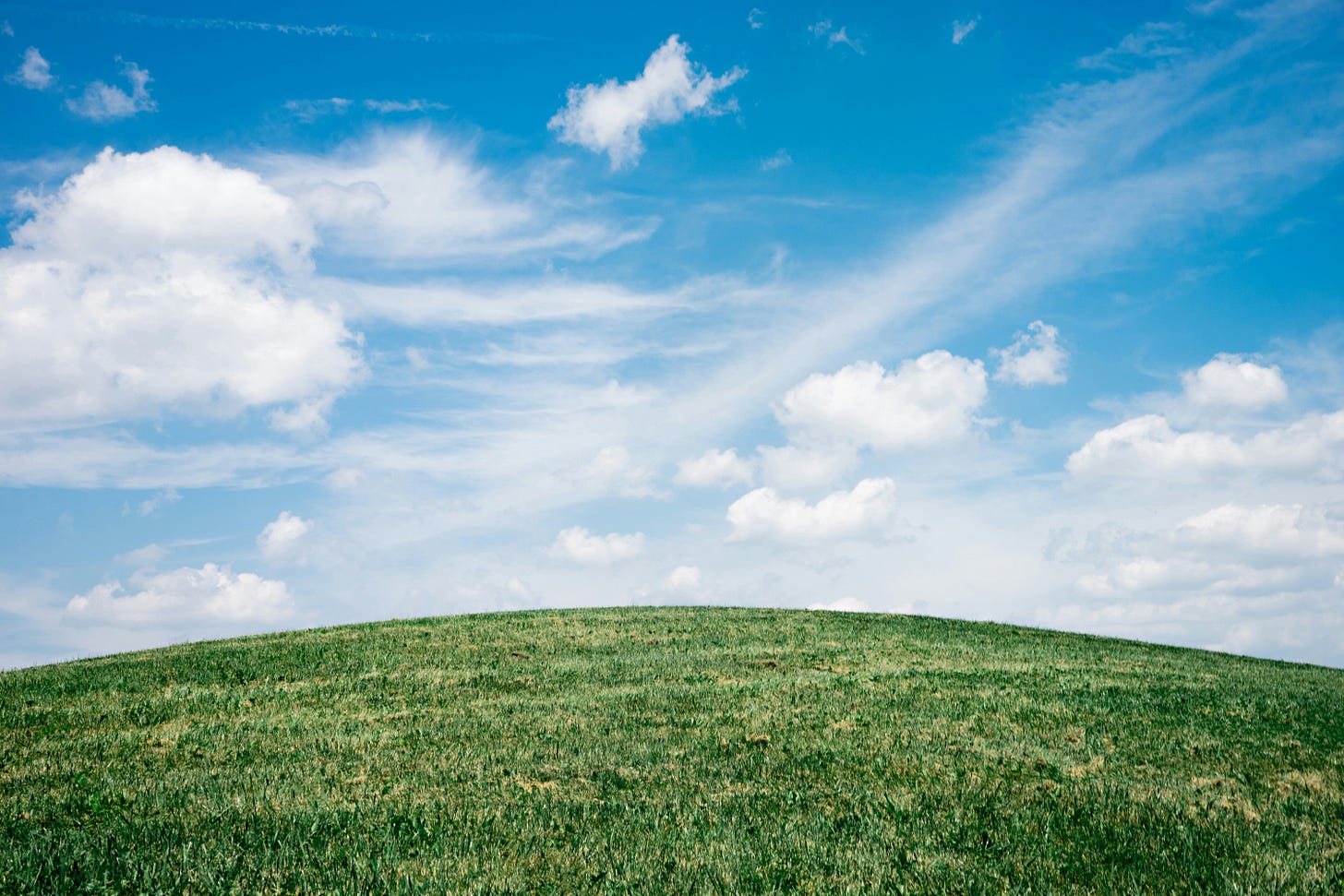 A serene landscape featuring a gentle grassy hill beneath a vibrant blue sky with scattered white clouds.
