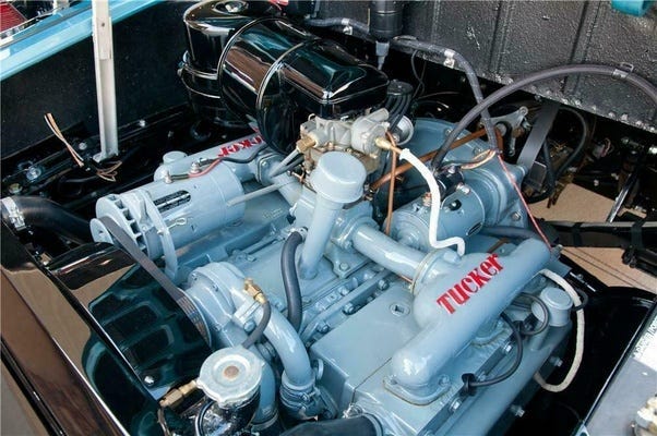 La imagen muestra una vista de cerca del motor de un coche antiguo, específicamente etiquetado como “TUCKER”. El motor parece estar bien mantenido y está instalado en el vehículo. Los componentes, incluyendo el filtro de aire, las tapas de las válvulas y varios tubos y conexiones, están pintados de un color azul claro uniforme con algunas partes en negro. La marca “TUCKER” se muestra de manera prominente en la tapa de las válvulas con letras rojas y un contorno blanco. La imagen muestra una vista de cerca del motor de un coche antiguo, específicamente etiquetado como “TUCKER”. El motor parece estar bien mantenido y está instalado en el vehículo. Los componentes, incluyendo el filtro de aire, las tapas de las válvulas y varios tubos y conexiones, están pintados de un color azul claro uniforme con algunas partes en negro. La marca “TUCKER” se muestra de manera prominente en la tapa de las válvulas con letras rojas y un contorno blanco.
