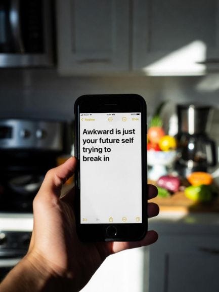 A hand holding a phone in a dark kitchen, the only light coming from the Notes app displaying the words: “Awkward is just your future self trying to break in.” A hand holding a phone in a dark kitchen, the only light coming from the Notes app displaying the words: “Awkward is just your future self trying to break in.”