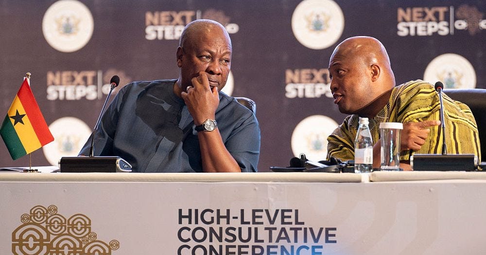 Two bald Black men sitting at a table with microphones, leaning towards each other in conversation Two bald Black men sitting at a table with microphones, leaning towards each other in conversation