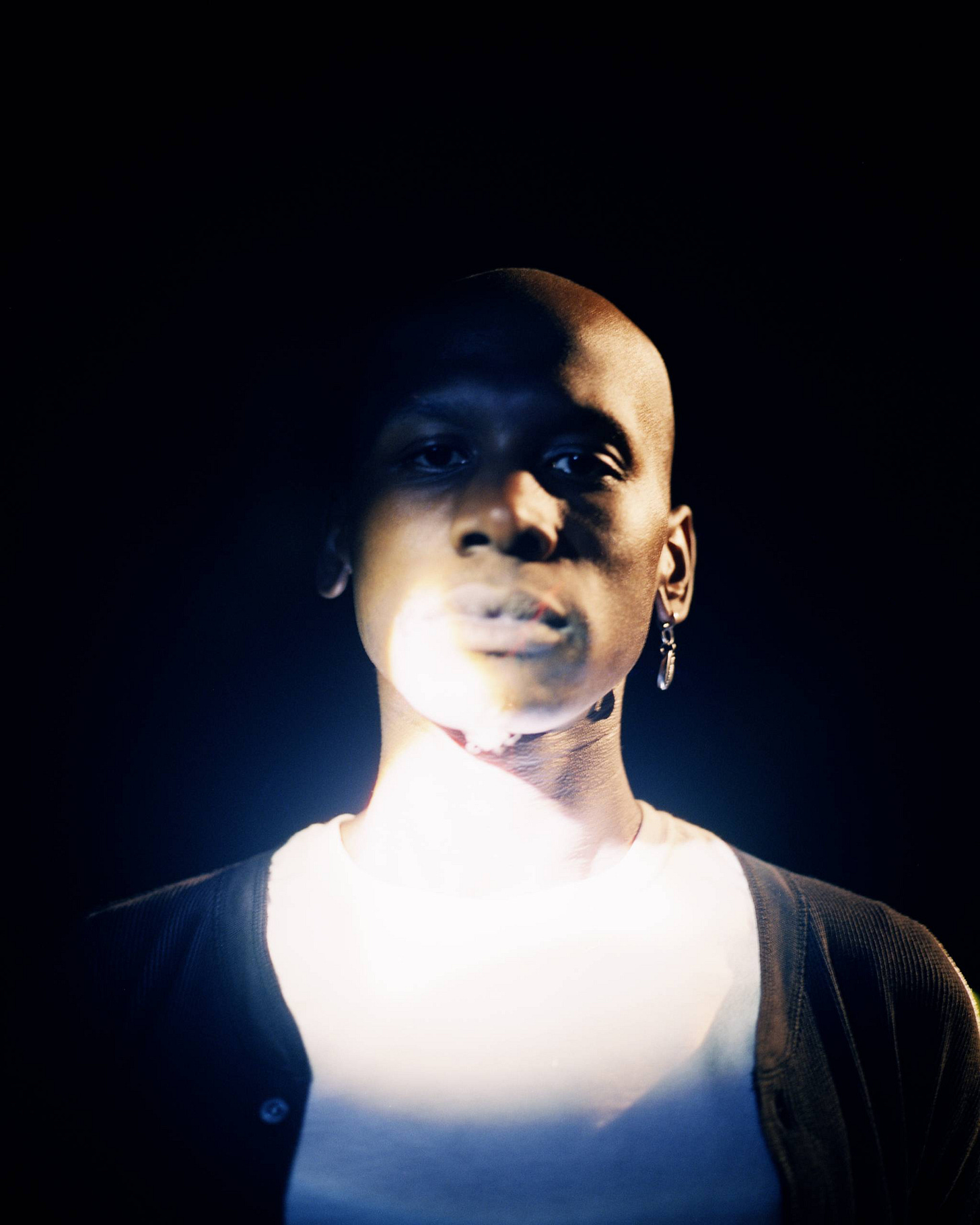 Person with shaved head wearing white shirt and dark cardigan, illuminated against black background with visible smoke.
