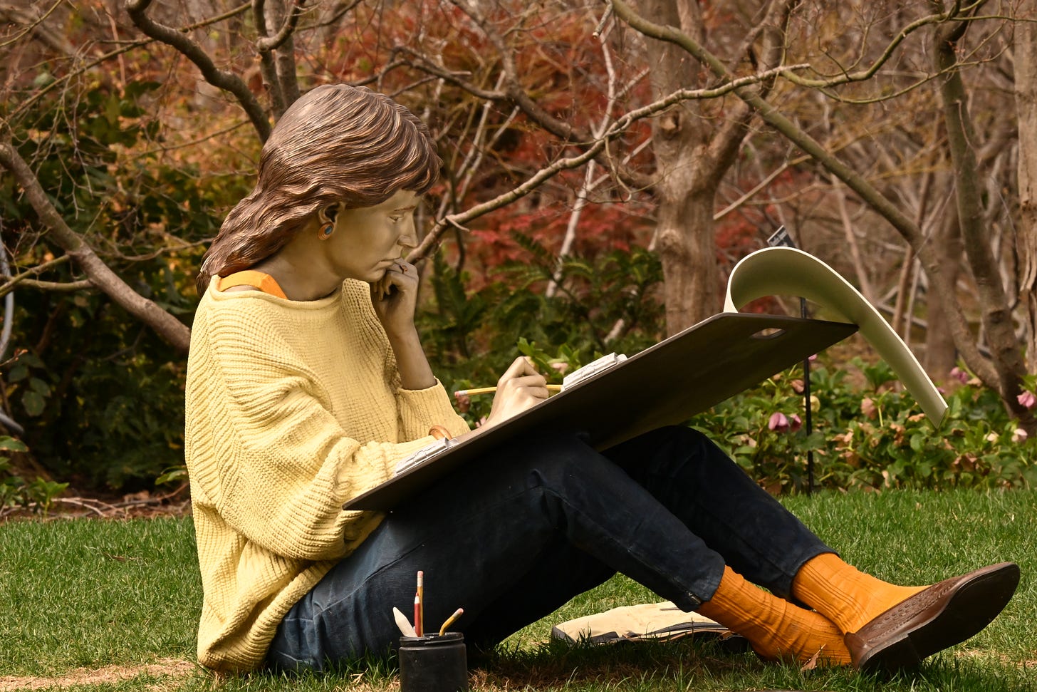 Metal sculpture of young woman, seated on the grass, sketching. She has light brown hair and wears a yellow sweater, jeans, and orange socks with brown leather flats. Her sketchpad is on her knees. Her left hand is at her chin in concentration. A cup with other drawing implements is in front of her. She faces the right of the frame.
