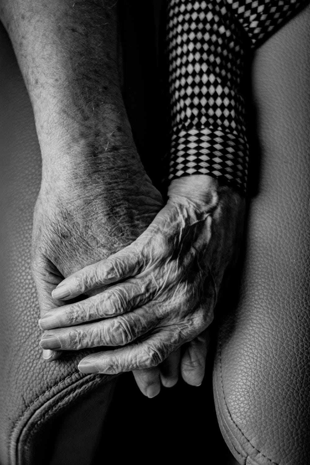 Two elderly hands, one resting on top of the other, on a leather sofa.