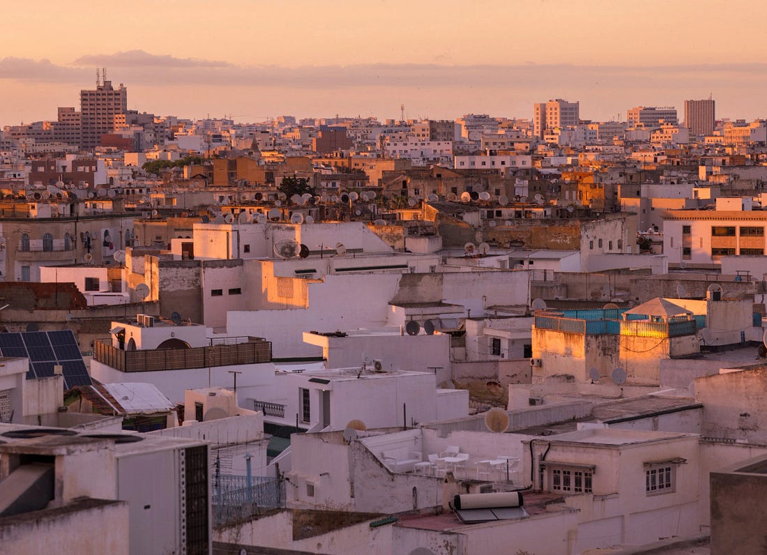 Rooftop view across central Tunis at sunset, showing dense white and tan buildings, satellite dishes, rooftop terraces, and modern high-rises rising beyond the older city.