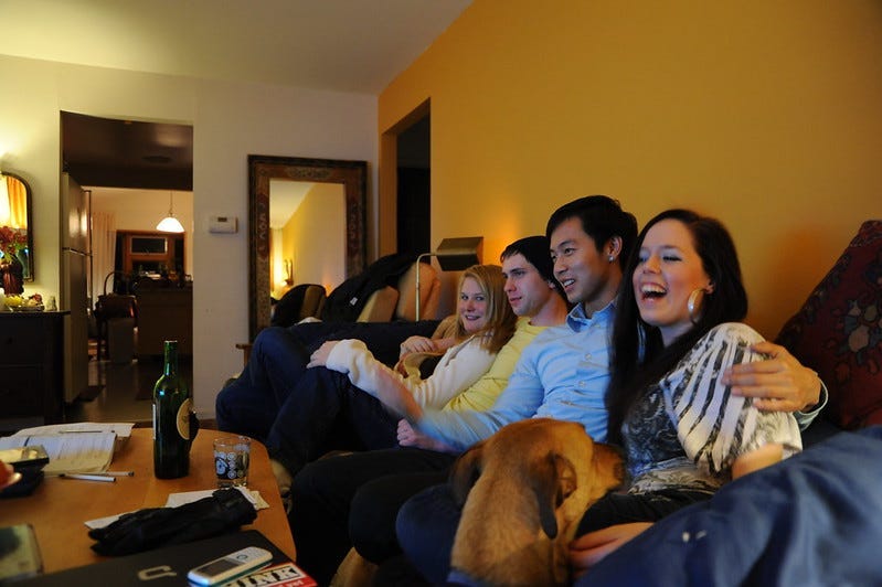 Four friends of different genders and ethnicities laughing on the couch while a dog tries to squeeze in.