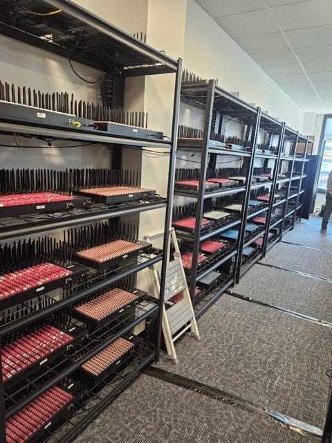 Multiple black SIM servers with numerous SIM card slots, some with red indicators, arranged on metal racks in a room. Several SIM servers and tangled cables scattered on a wooden floor in a room with a window, dark curtains, and a door. No watermarks from platforms like Instagram, TikTok, or Xiaohongshu are present.