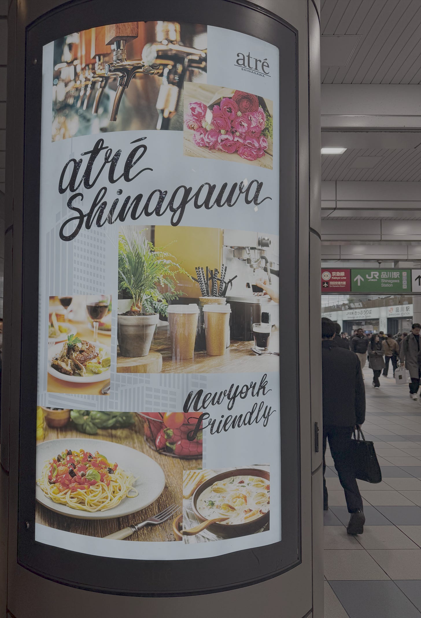 A curved digital advertisement for Atré Shinagawa, a shopping complex located inside JR Shinagawa Station in Tokyo. The display showcases the venue's dining and lifestyle offerings — craft beer taps, fresh flowers, gourmet dishes, coffee, and pasta — under the tagline "New York Friendly." Commuters pass by in the station concourse, with signage for the Keikyū Line and JR Shinagawa Station visible in the background.