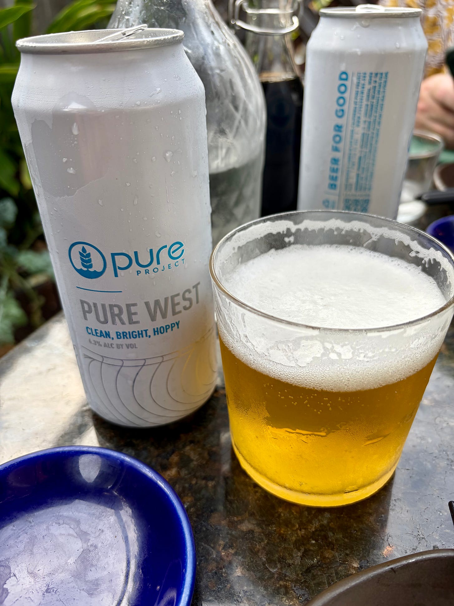 A close-up shot of a chilled white can of Pure Project "Pure West" West Coast IPA covered in condensation. Next to the can is a short, wide glass filled with a golden, frothy beer with a thick white head, sitting on a dark granite table top.