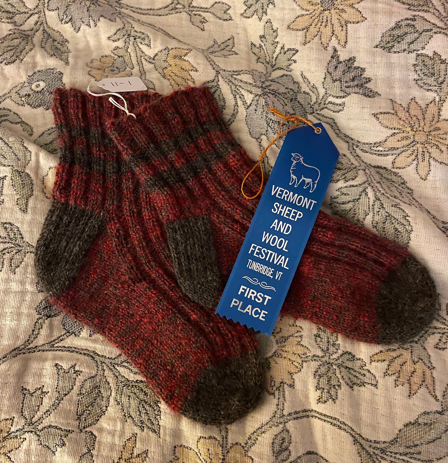 Dark Grey and Red marled hand knit socks with dark grey stripes, heel and toes sit atop a floral blanket. There is a blue award ribbon that reads "Vermont Sheep and Wool Fest, Tunbridge, VT, First Place"