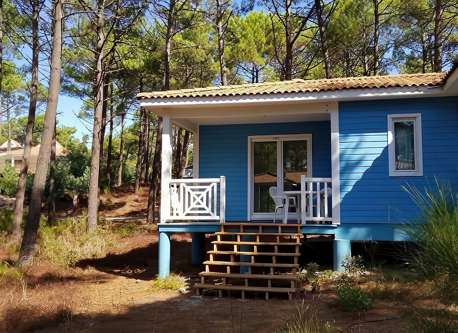 A blue wooden chalet with white railing and covered terrace, elevated on stilts above the pine-needle floor of La Jenny's maritime forest, Le Porge, Gironde.