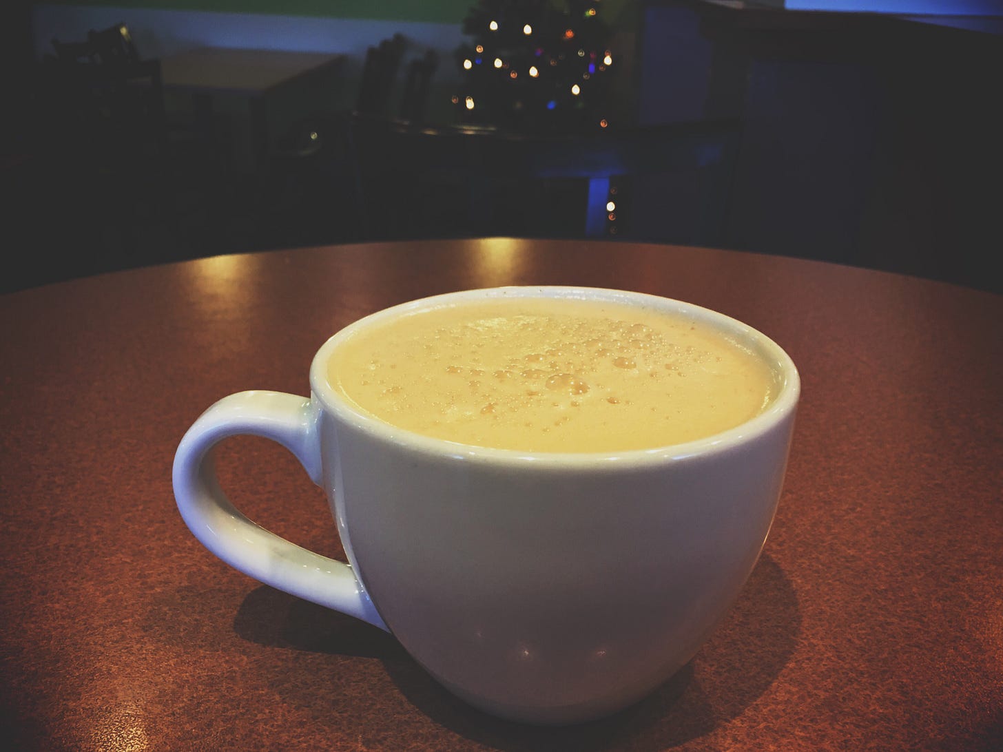 A mug of eggnog on a wooden table in a dimly lit bar, with soft holiday lights in the background.
