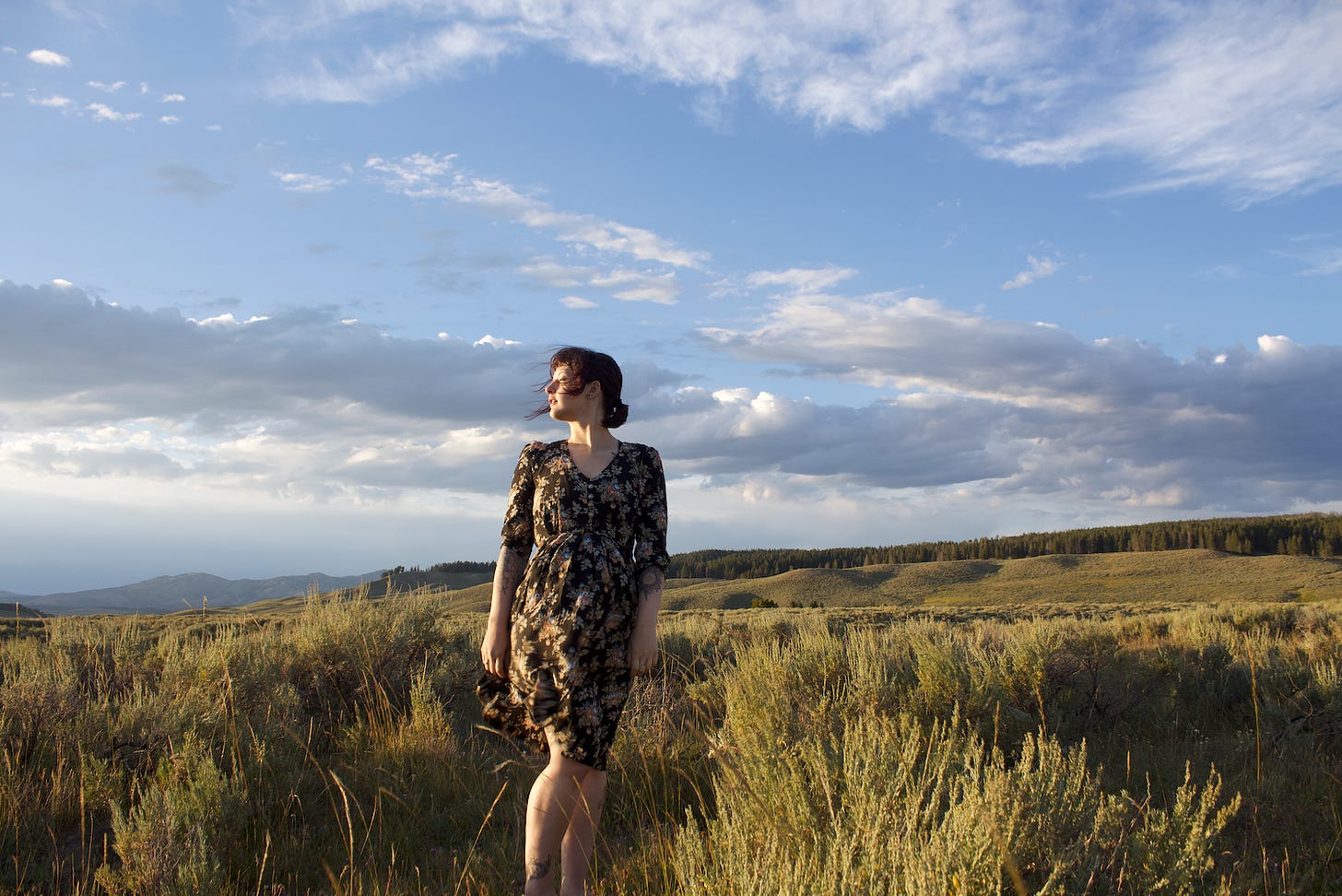 A person in a dark floral dress stands in a wide, sunlit field of sagebrush and grasses at golden hour, looking toward the horizon under a sky of drifting clouds.