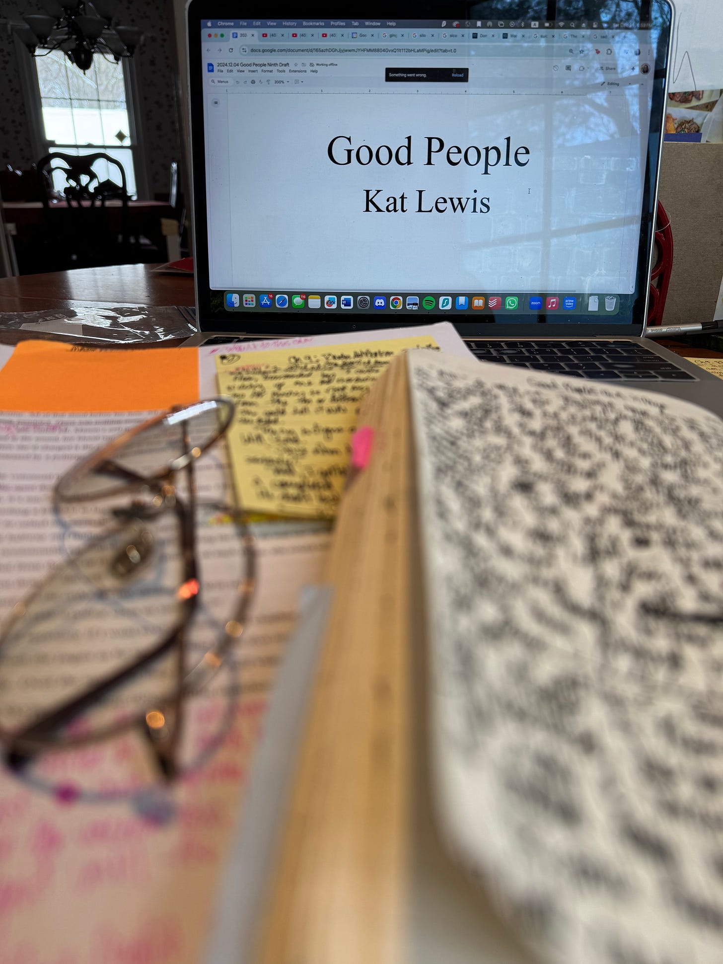 A photo of Kat’s monitor opened to the title page of Good People. Her printed manuscript and notebook are opened in front of her computer, each page filled with messy handwriting.
