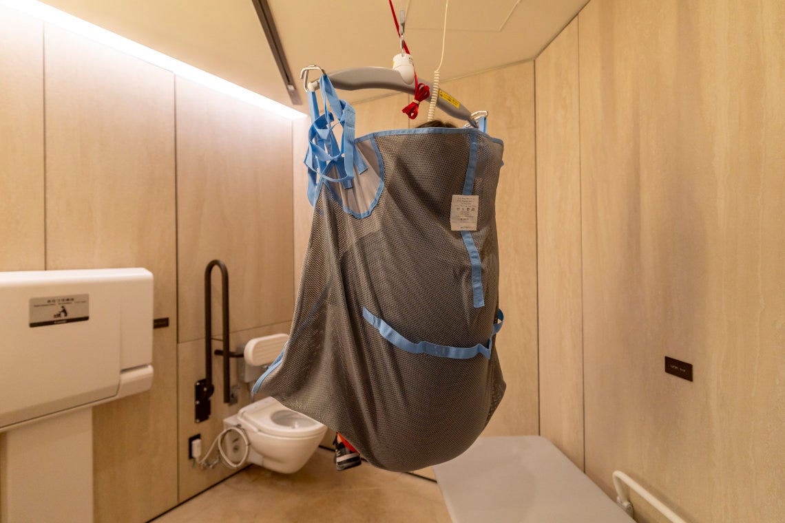Ceiling lift with a gray and blue sling suspended above an accessible restroom with beige walls.