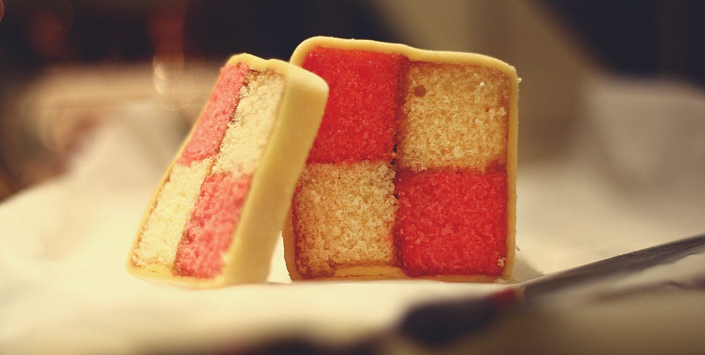 A close-up view of two slices of Battenberg cake, revealing the classic pink and yellow checkered sponge pattern held together with jam and encased in a thin layer of pale yellow marzipan. One slice leans against the other on a white surface, with a blurred background suggesting an indoor setting, possibly a table with soft lighting. A close-up view of two slices of Battenberg cake, revealing the classic pink and yellow checkered sponge pattern held together with jam and encased in a thin layer of pale yellow marzipan. One slice leans against the other on a white surface, with a blurred background suggesting an indoor setting, possibly a table with soft lighting.
