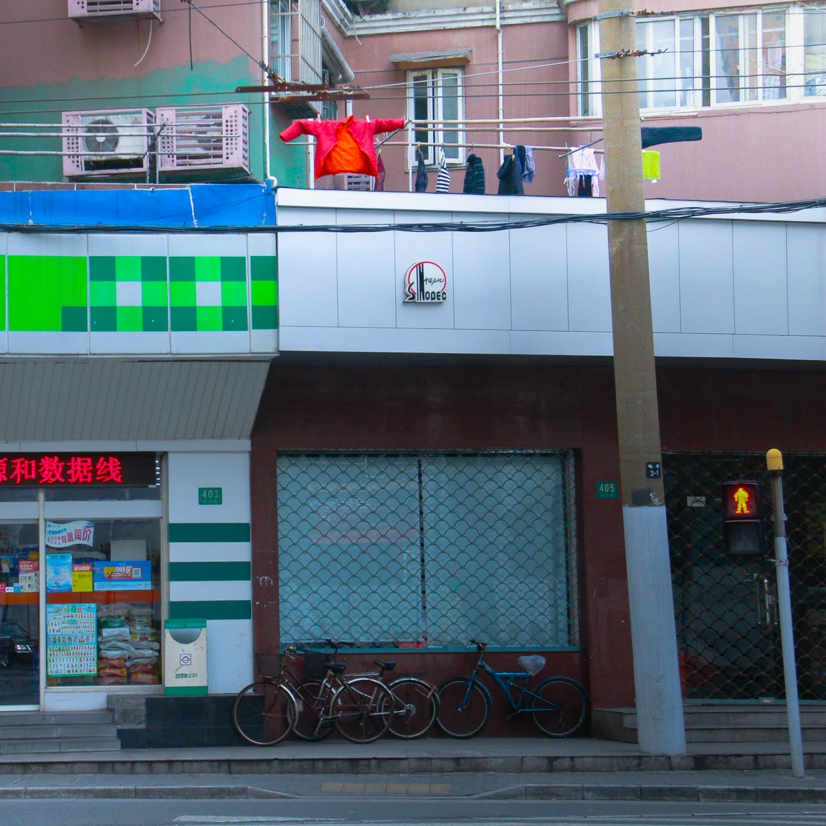 A vivid red puffy coat outstretched over a storefront awning. Bicycle-lined sidewalk and signage mark this quintessential Shanghai street corner.