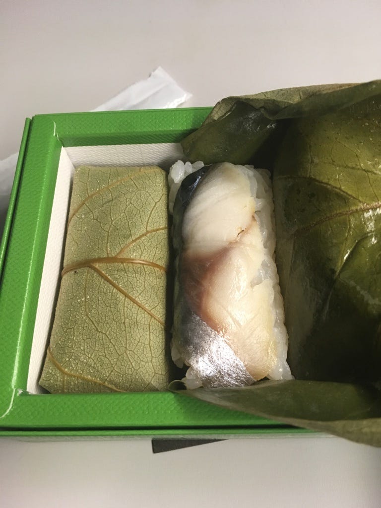 2 rectangular pressed sushi pieces wrapped in green persimmon leaves. The left one is still wrapped fully. The right one is open, showing a silver and white mackerel slice on top of white rice. 2 rectangular pressed sushi pieces wrapped in green persimmon leaves. The left one is still wrapped fully. The right one is open, showing a silver and white mackerel slice on top of white rice.