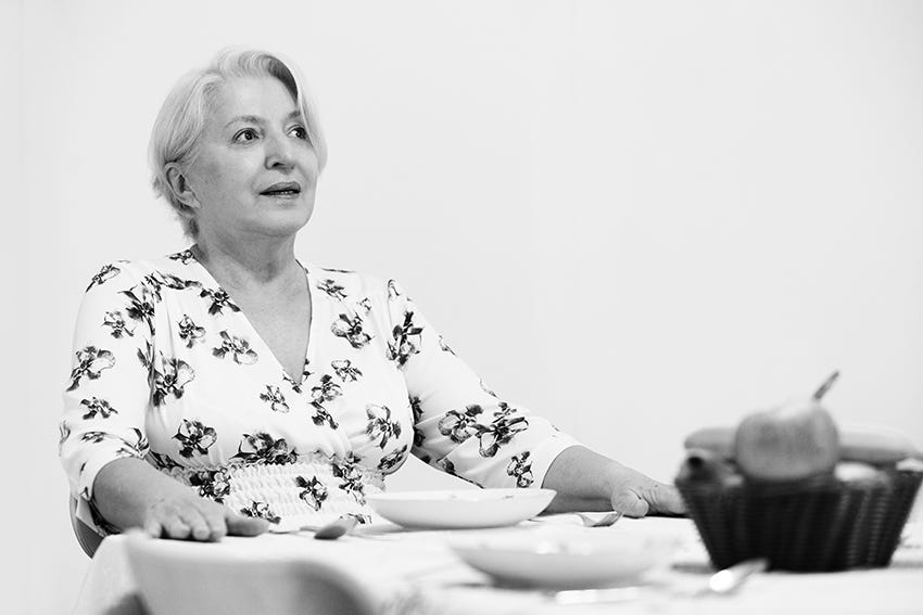 A black and white image of Sadika sitting at table on her own. There is a bowl of fruit on the table and two place settings.