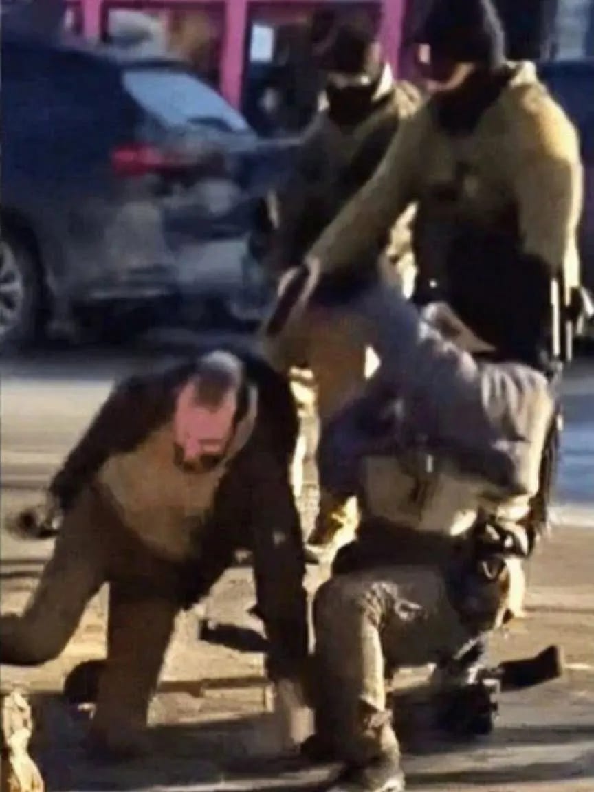 A disturbing image showing Alex Pretti in the moment before his murder. His left hand is on the pavement, his right hand holds his phone. He is on his knees while an agent of Customs and Border Patrol is pointing a gun at Pretti's head from point-blank distance.