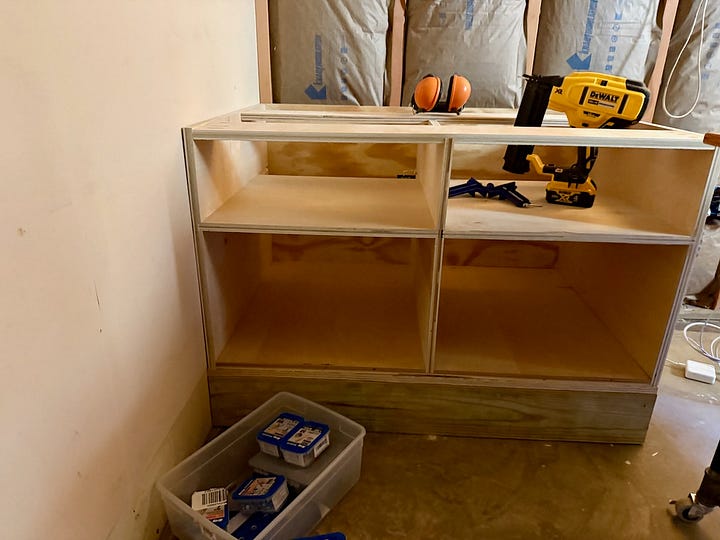 Two photos of basement cabinetry construction. The first shows a close-up of a plywood cabinet box with tools on top and storage bins on the floor. The second shows a wider view with two cabinet bases installed against an insulated wall, wiring exposed overhead.