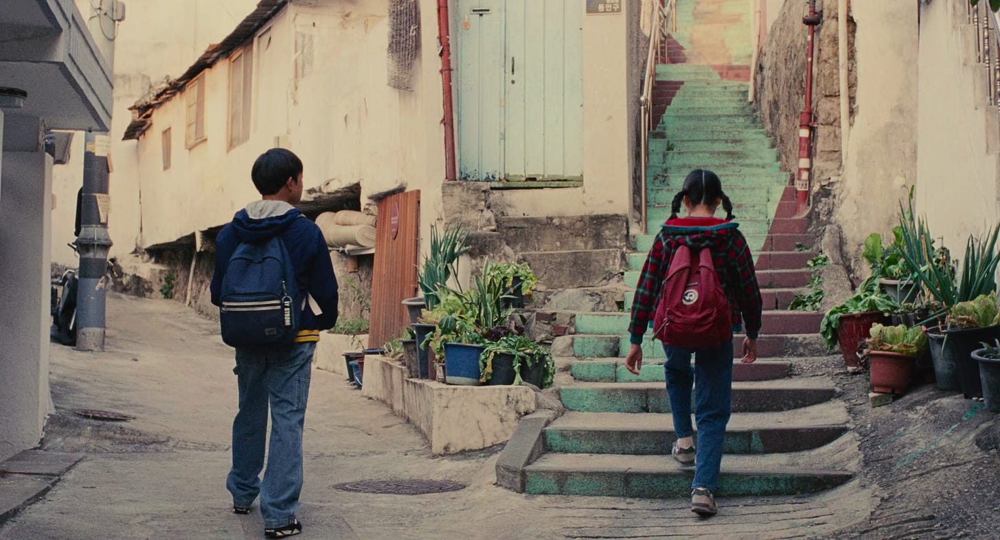 Dois jovens de mochila vistos de costas - o menino Hae Sung segue pela rua à esquerda enquanto a menina Na Young sobe uma escadaria turquesa à direita, simbolizando os caminhos que se separam na primeira despedida.