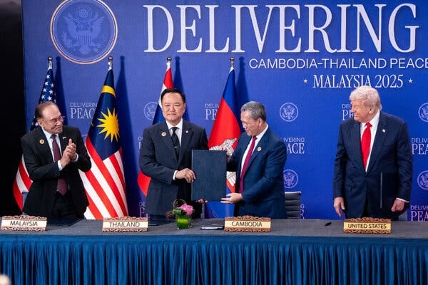 Two men hold a large blue binder as they stand behind a long table. Behind them are different national flags. 