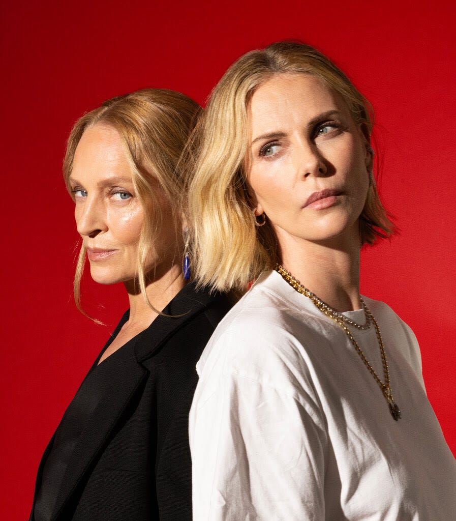 Two women with blonde hair stand back-to-back against a bright red background. One wears a black blazer and looks at the camera, the other wears a white top and gold chain, gazing to the side.