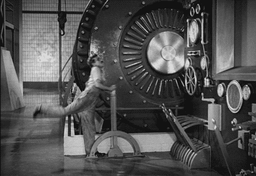 Gif in bianco e nero da "Tempi moderni", con Charlie Chaplin vestito con una salopette da operaio mentre si appoggia a una leva gigante con un passo da danzatore. Sullo fondo una gigante ruota dentata con dei meccanismi.