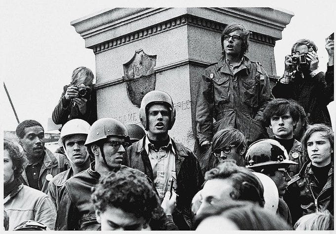 A black-and-white photo of a crowd gathered around a stone monument. People wear helmets and casual clothing, some holding cameras. The scene depicts a public demonstration with individuals standing closely together.
