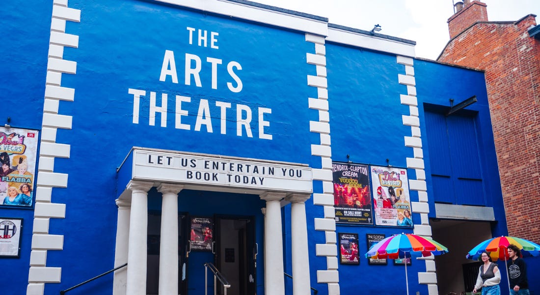A blue and white theatre with colourful umbrellas and people walking in front of it A blue and white theatre with colourful umbrellas and people walking in front of it