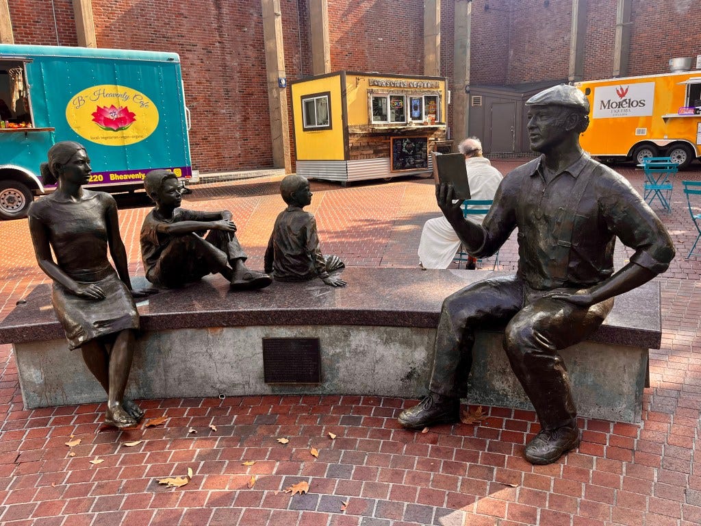 A bronze statue of Ken Kesey seated on a bench and reading to three of his grandchildren, a teenage girl and two young boys. He hold a book aloft and rests a hand on his thigh. On his head, a flat cap. Foodtrucks are parked in the brick-laid plaza, a part of Eugene, Oregon.