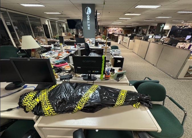 A body-shaped package wrapped in garbage bags & bound in crime scene-tape lies on a desk in a newsroom.
