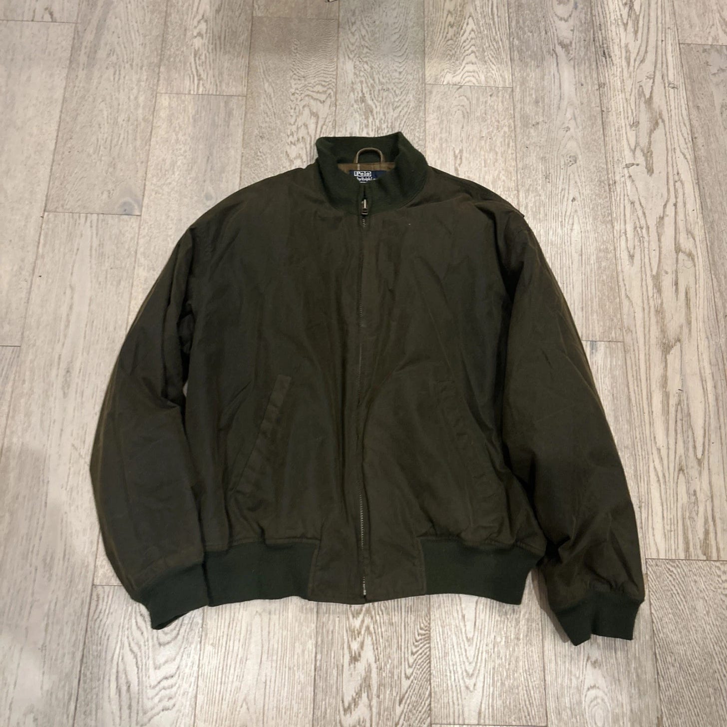 Olive green vintage Polo Ralph Lauren men’s waxed cotton oilcloth bomber jacket with collar and ribbed cuffs, laid flat on light wood floor. Olive green vintage Polo Ralph Lauren men’s waxed cotton oilcloth bomber jacket with collar and ribbed cuffs, laid flat on light wood floor.