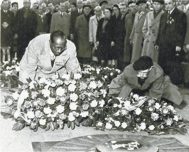 Black man in suit with medals and white man in beret lay large wreaths, surrounded by crowd