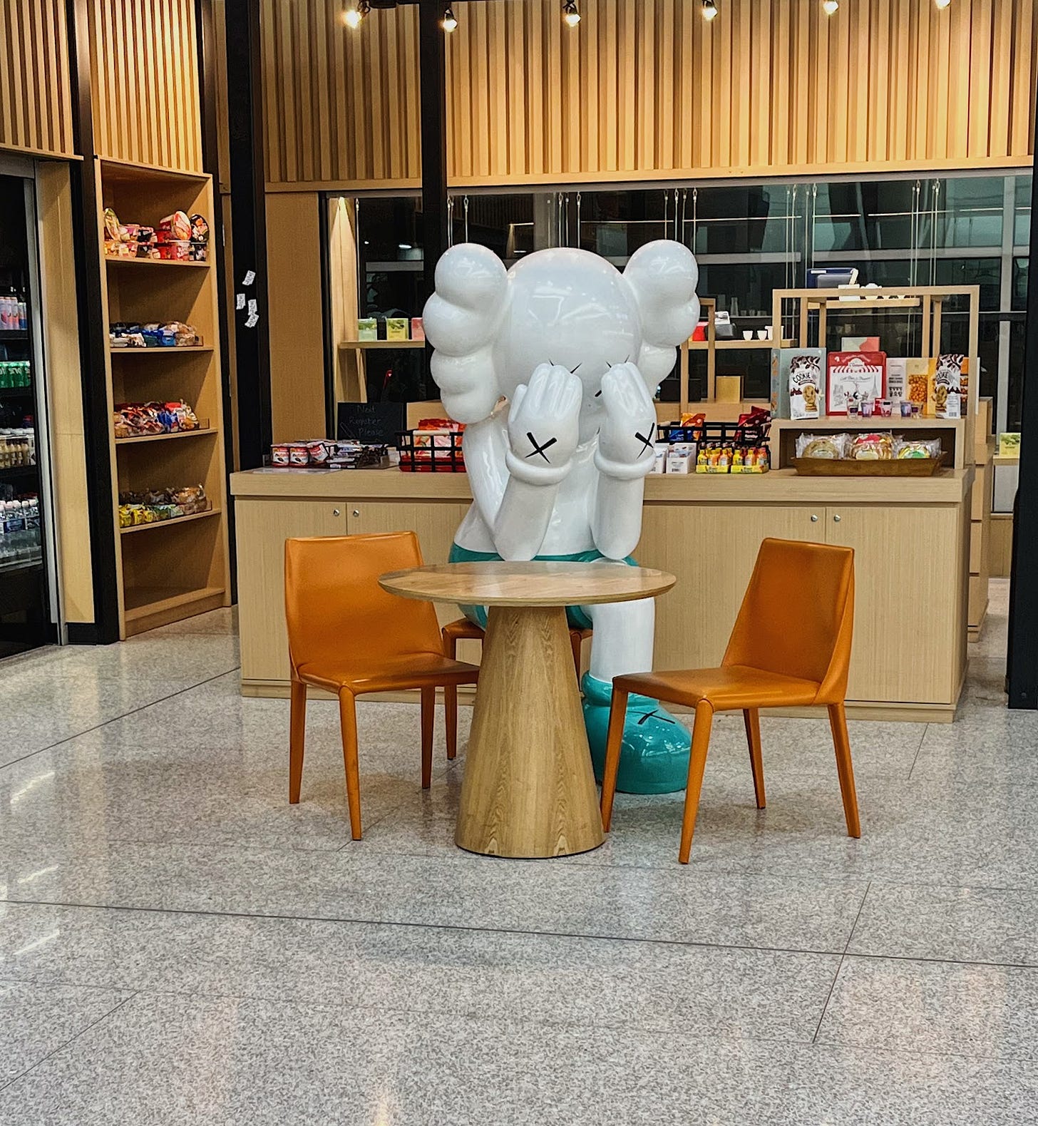 A giant white statue of a cartoonish man with mickey like gloves looking down into its hands at a cafe table