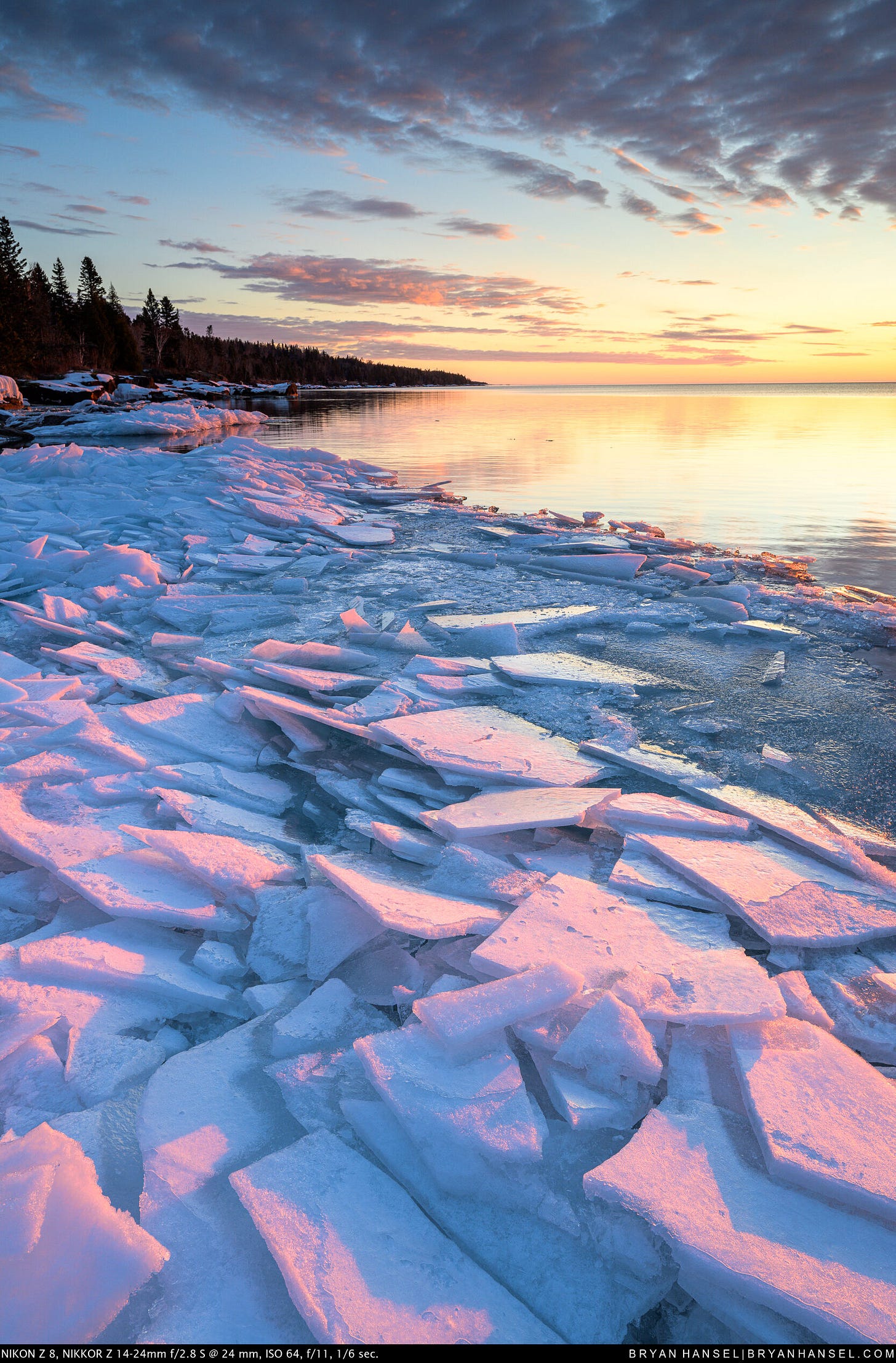 Ice plates painted pink and blue from sun and shadow lead out to open water under a sunrise on Lake Superior.