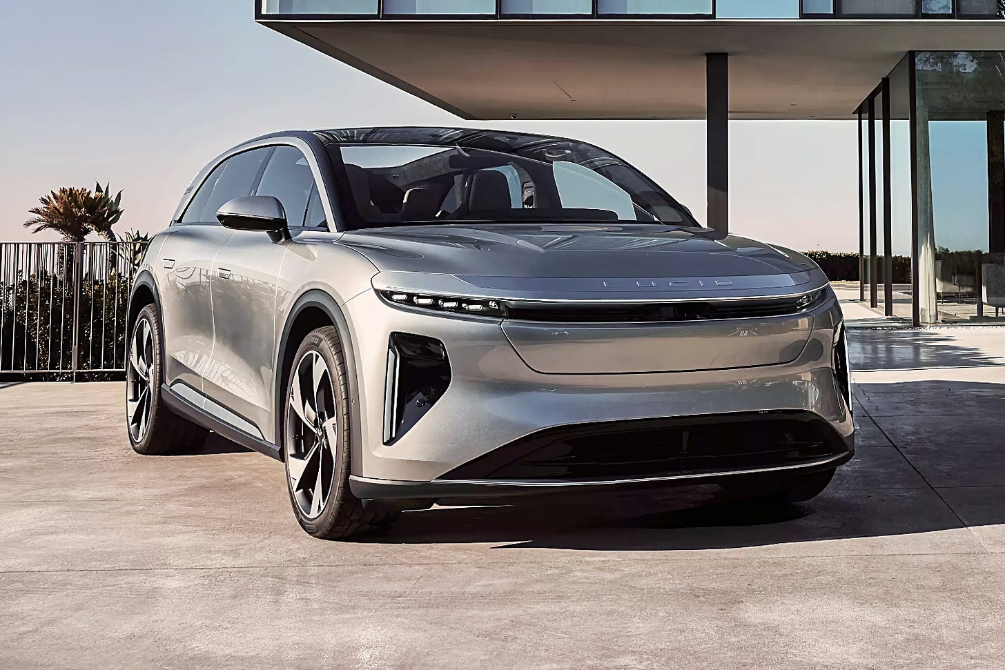 A silver Lucid Gravity electric SUV is parked on a driveway outside of a modern home with lots of windows.