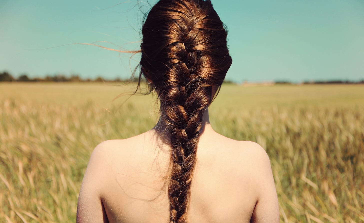 A woman with long brown hair in a braid stands nude from the waist up, facing a field of tall grass under a clear blue sky.
